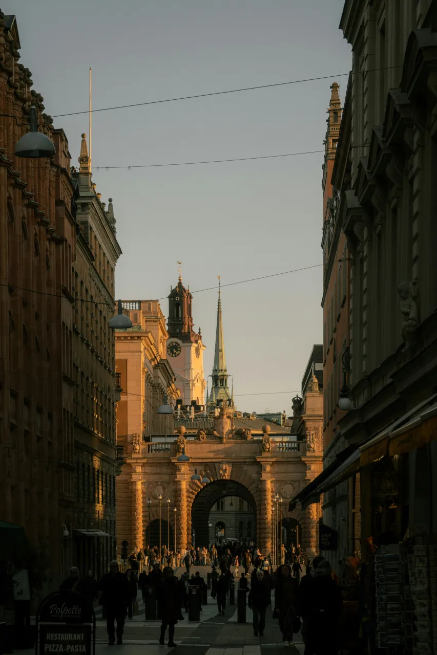Gamla Stan street in Stockholm with Riddarholmen Church tower at sunset, historic old town architecture and people walking